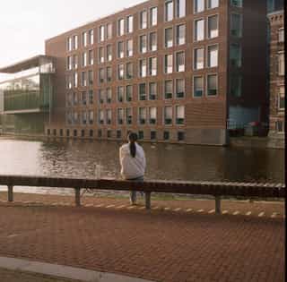 Person seen from behind, sitting on a bench by a canal facing a brick building